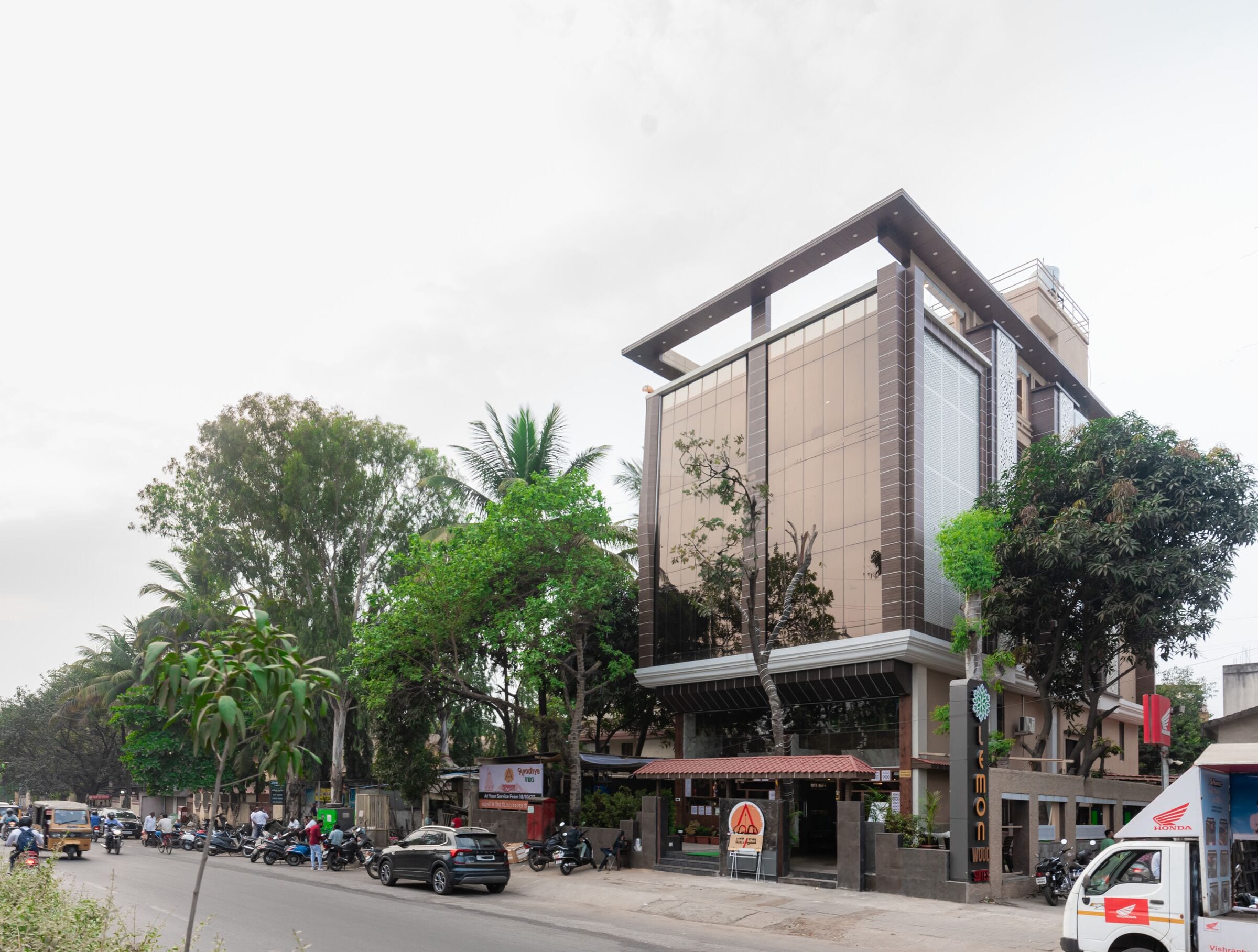 Lemonwood Suites by day: a tall walnut-brown facade with a square rooftop frame, behind palms and street trees